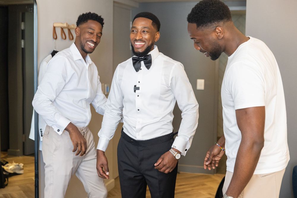 Candid moment of three friends laughing together during formal wedding preparations in a bright room.