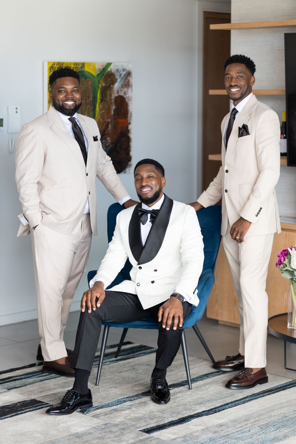 Three groomsmen pose in elegant cream and white tuxedos with black accents in a modern indoor setting.