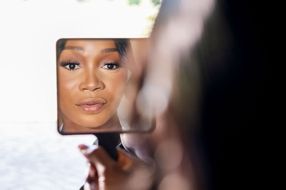 A reflection in a handheld mirror shows someone's face with soft natural makeup and a gentle smile.