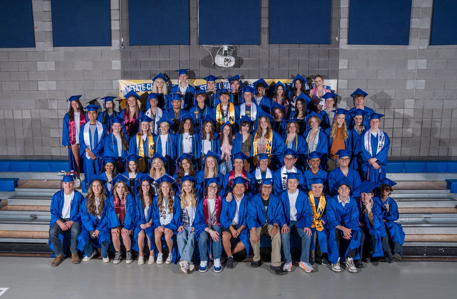 Large group of graduates in bright blue academic regalia seated on bleachers for formal class photo.