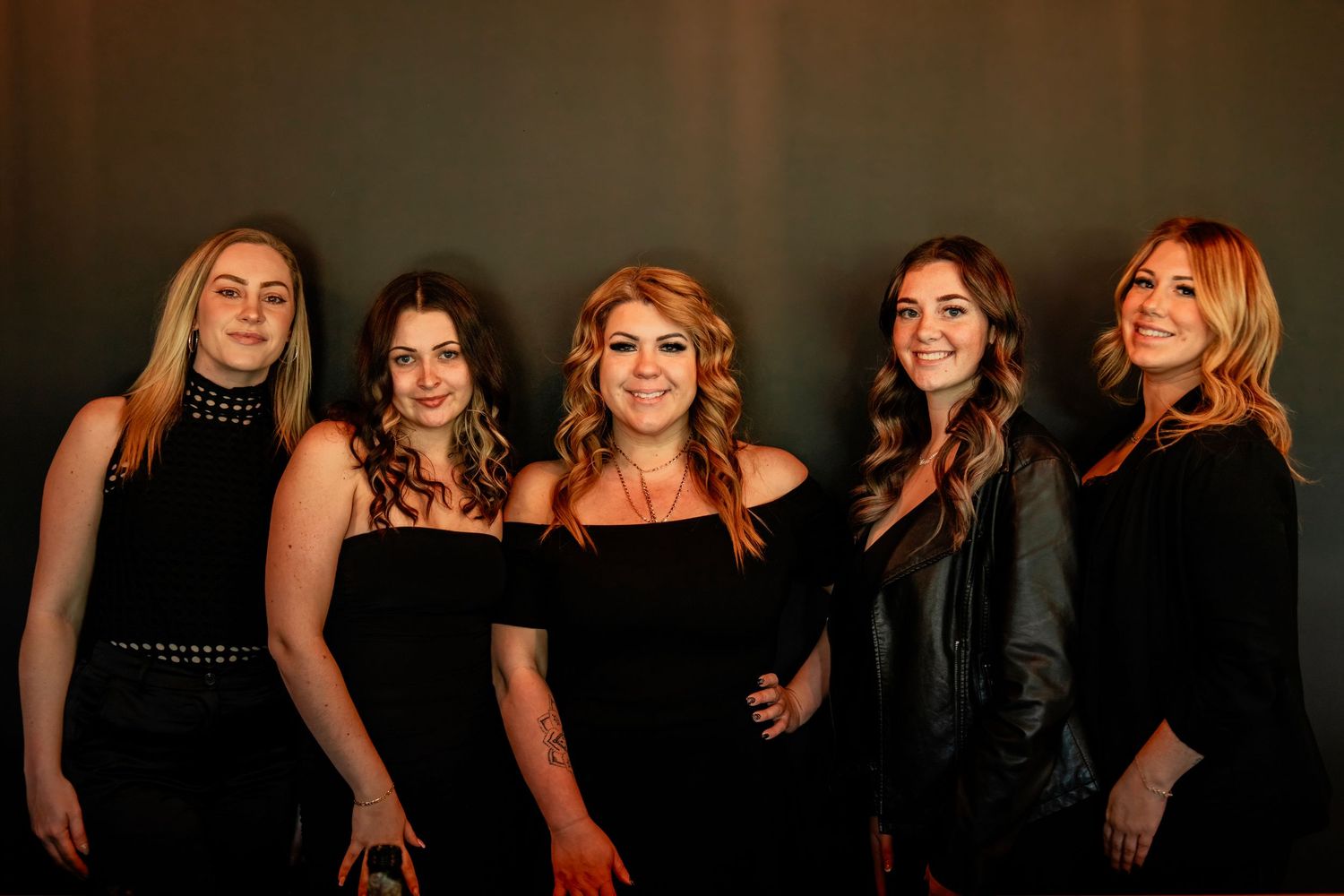 Group portrait of five women in elegant black attire against dark background.