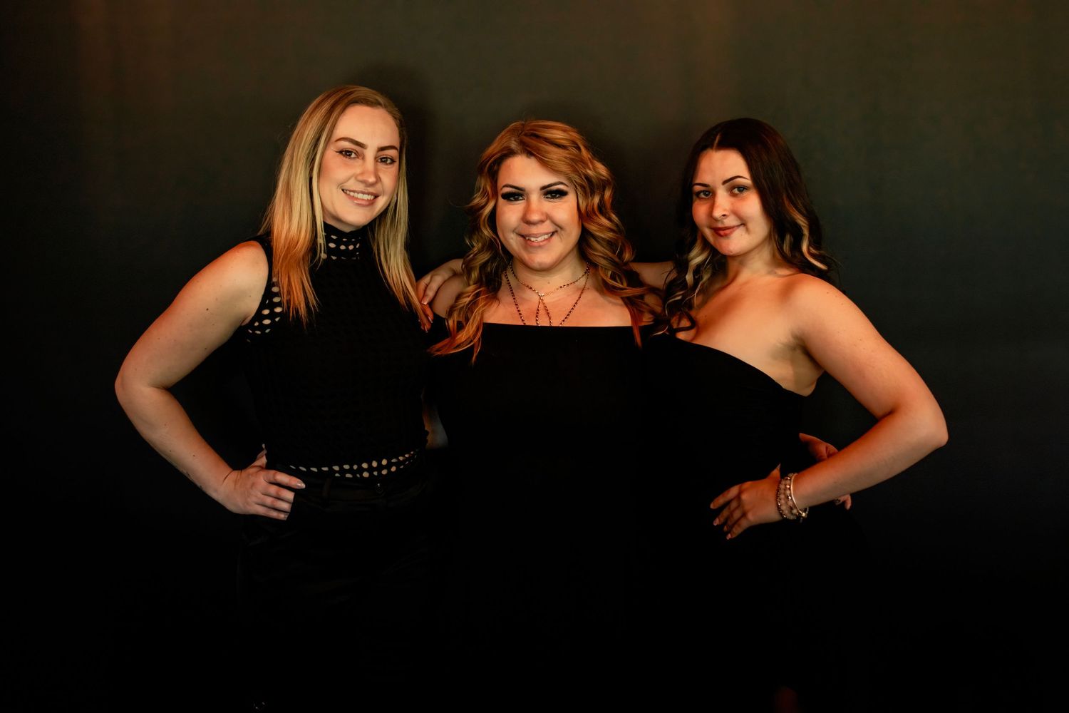 Three women in black formal wear pose together for dramatic studio portrait.