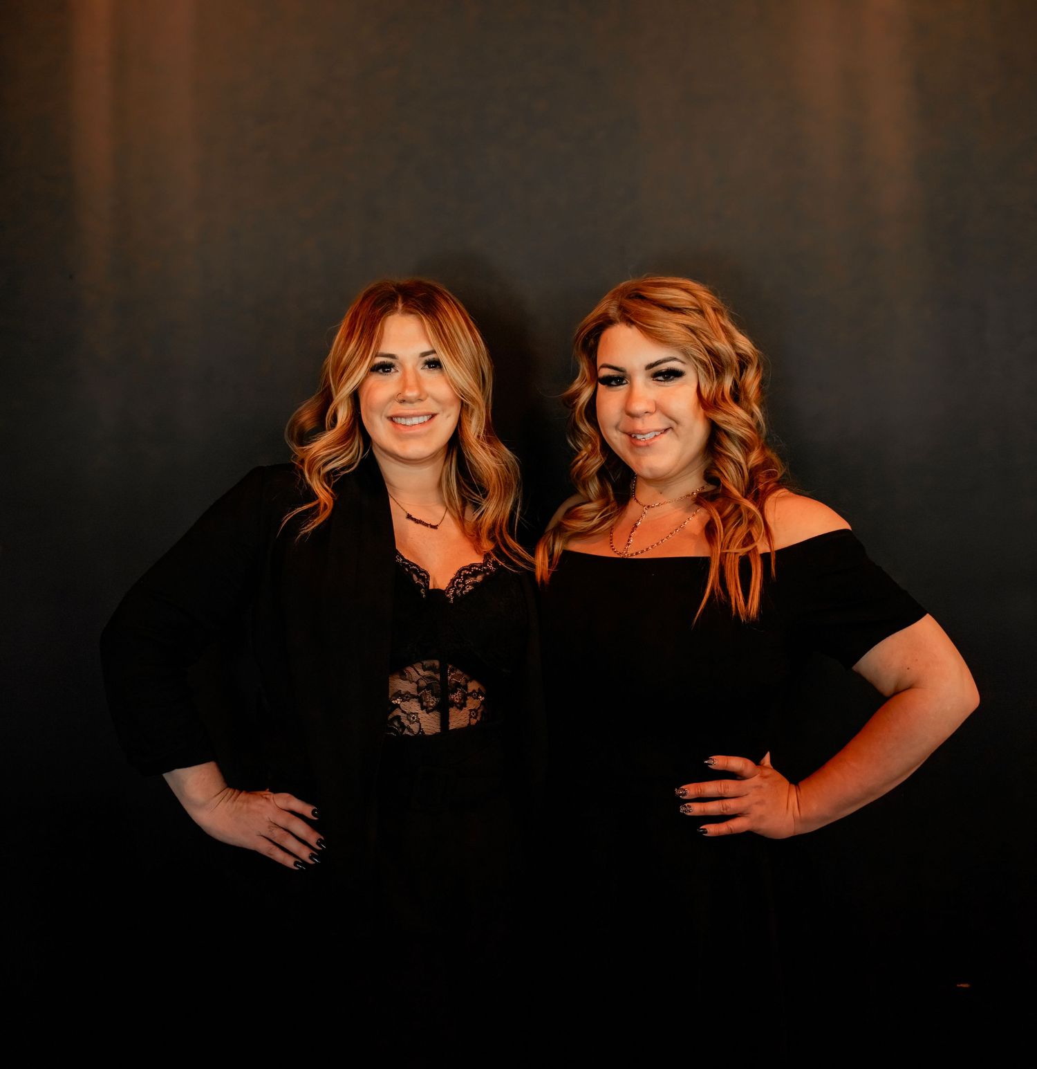 Two women in black outfits pose together for dramatic studio photograph.