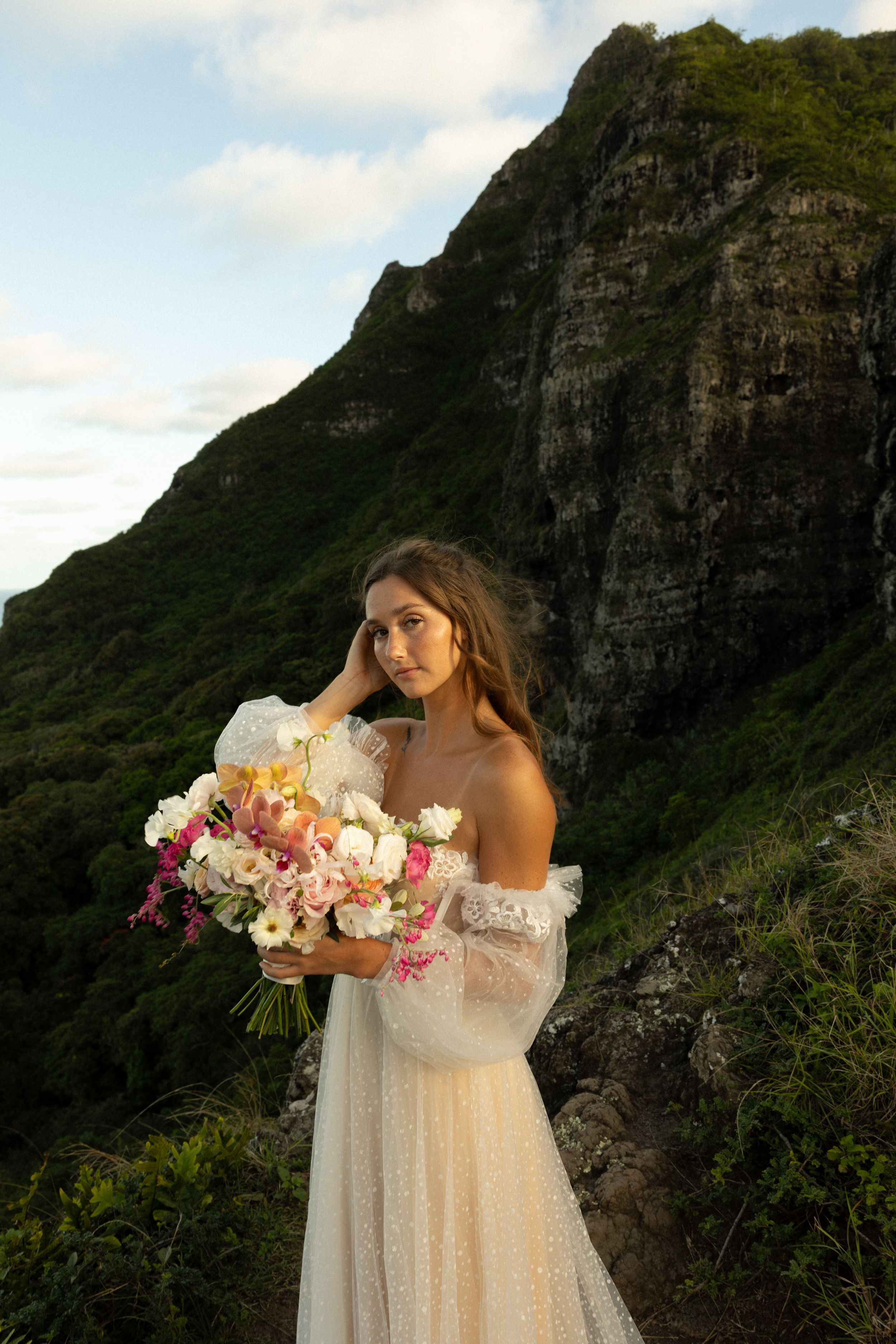 A bride in an off-shoulder wedding dress holds a pink and white bouquet against a dramatic mountain backdrop.