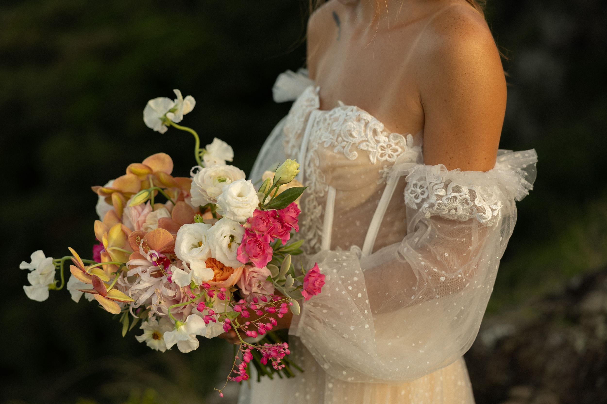 A delicate bridal bouquet of pink, orange and white flowers held against a white lace gown with off-shoulder sleeves.