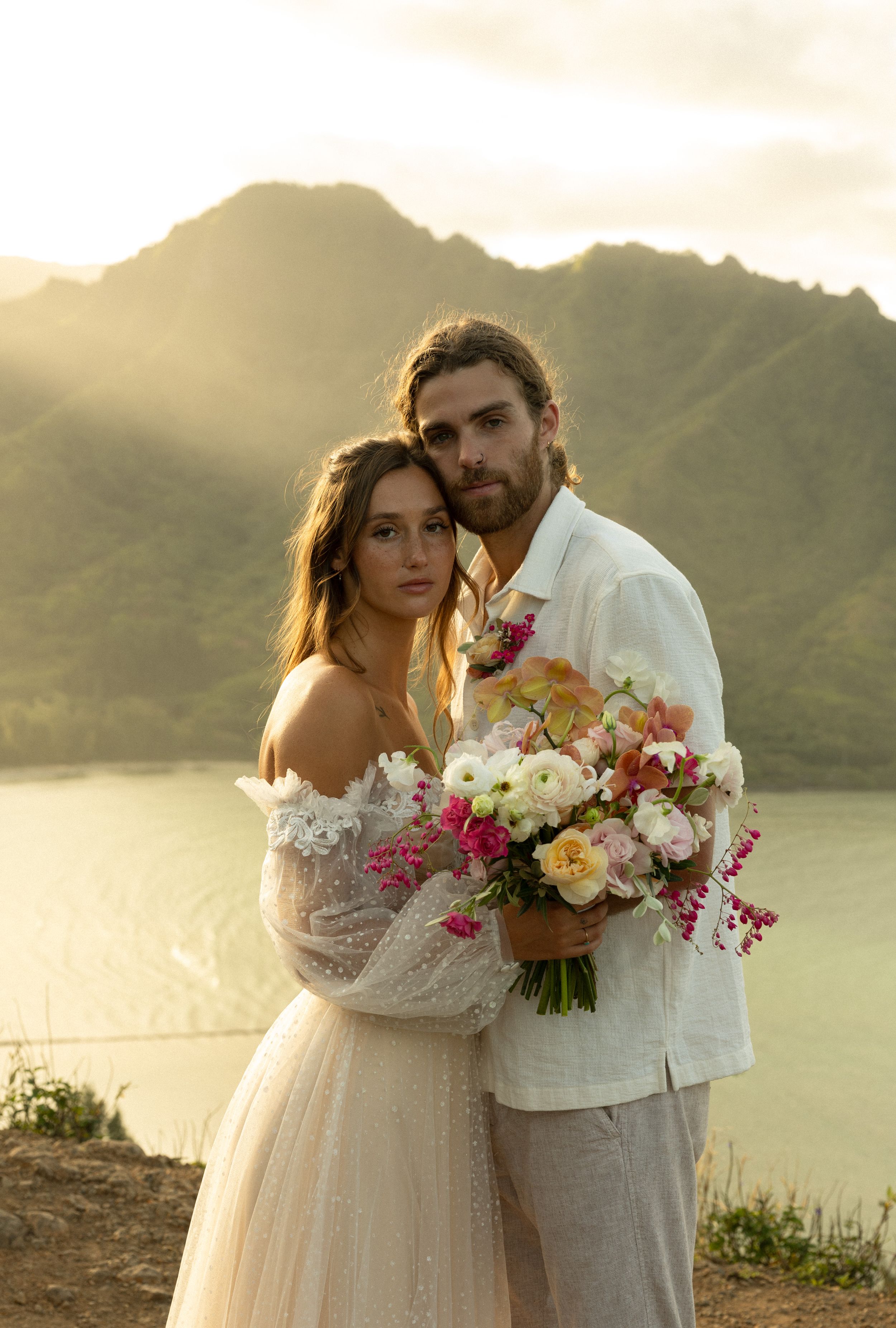 A romantic couple poses at sunset against mountain scenery with a stunning floral bouquet in warm golden light.