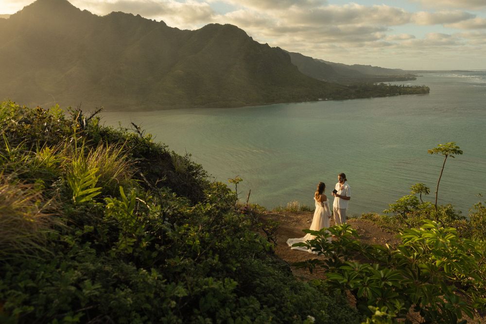 Scenic sunset view overlooking Oahu's Kaneohe Bay as two figures stand on hiking trail surrounded by lush vegetation.