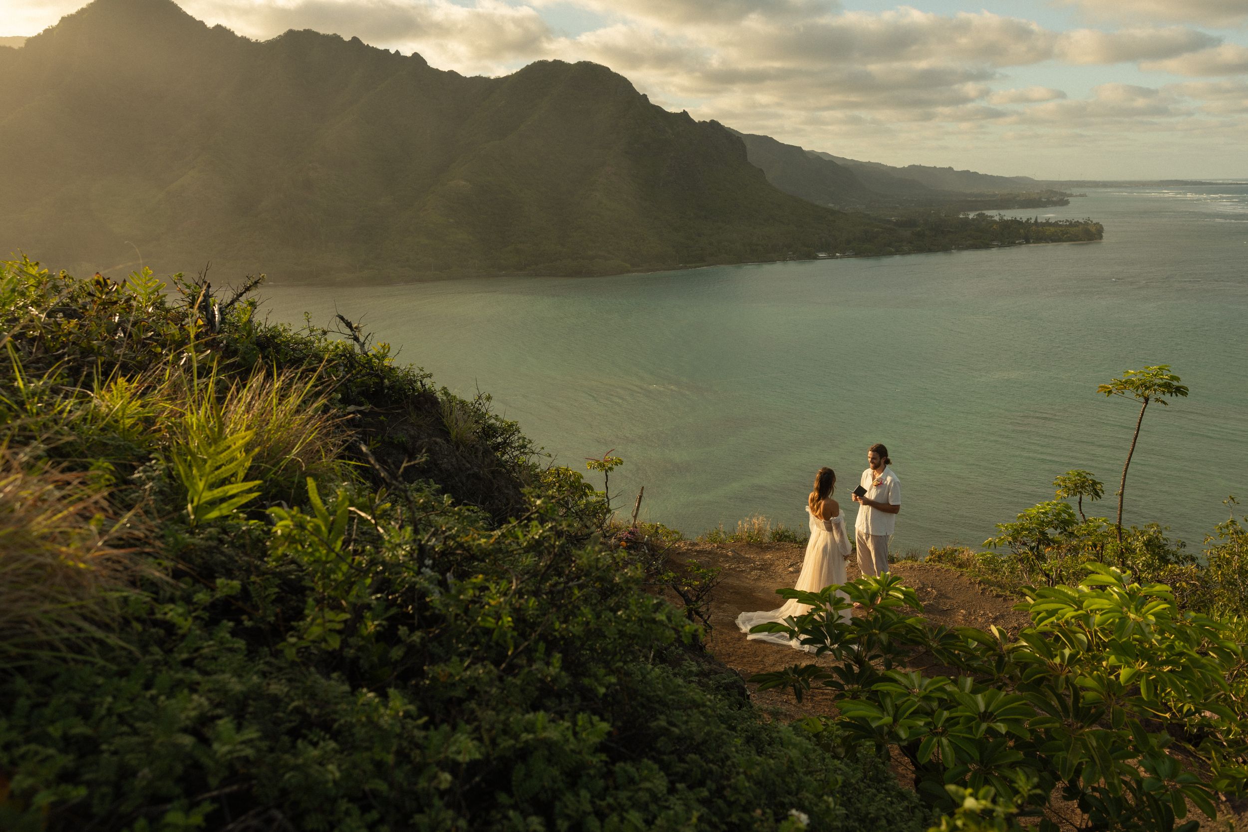 Scenic sunset view overlooking Oahu's Kaneohe Bay as two figures stand on hiking trail surrounded by lush vegetation.
