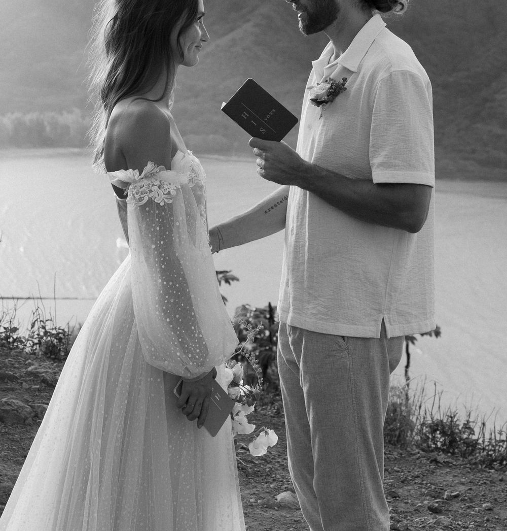 A couple exchanges vows during their mountain top ceremony with water views in the background.