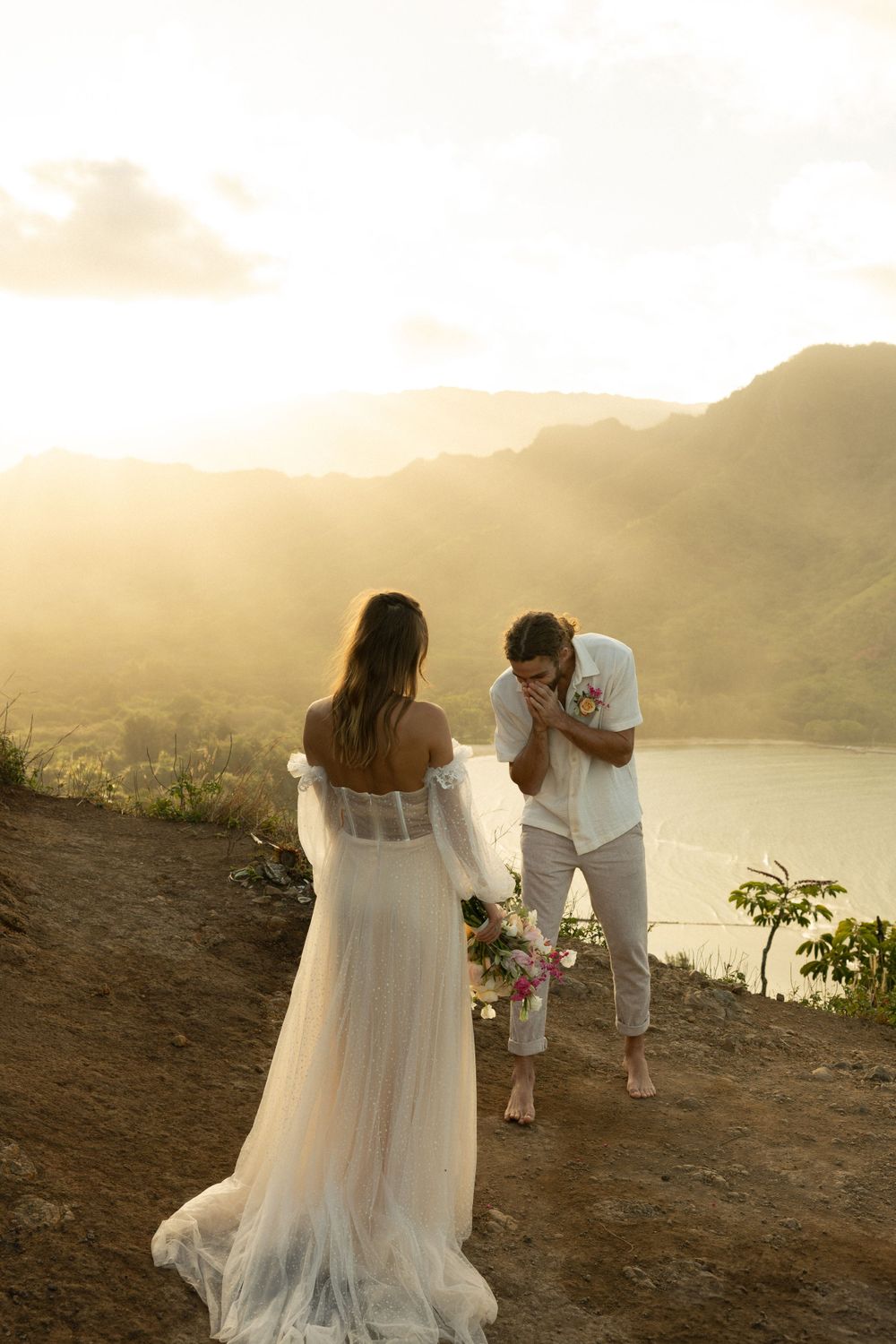 A couple shares a romantic moment at sunset overlooking mountains and water, with the bride in a flowing white dress.