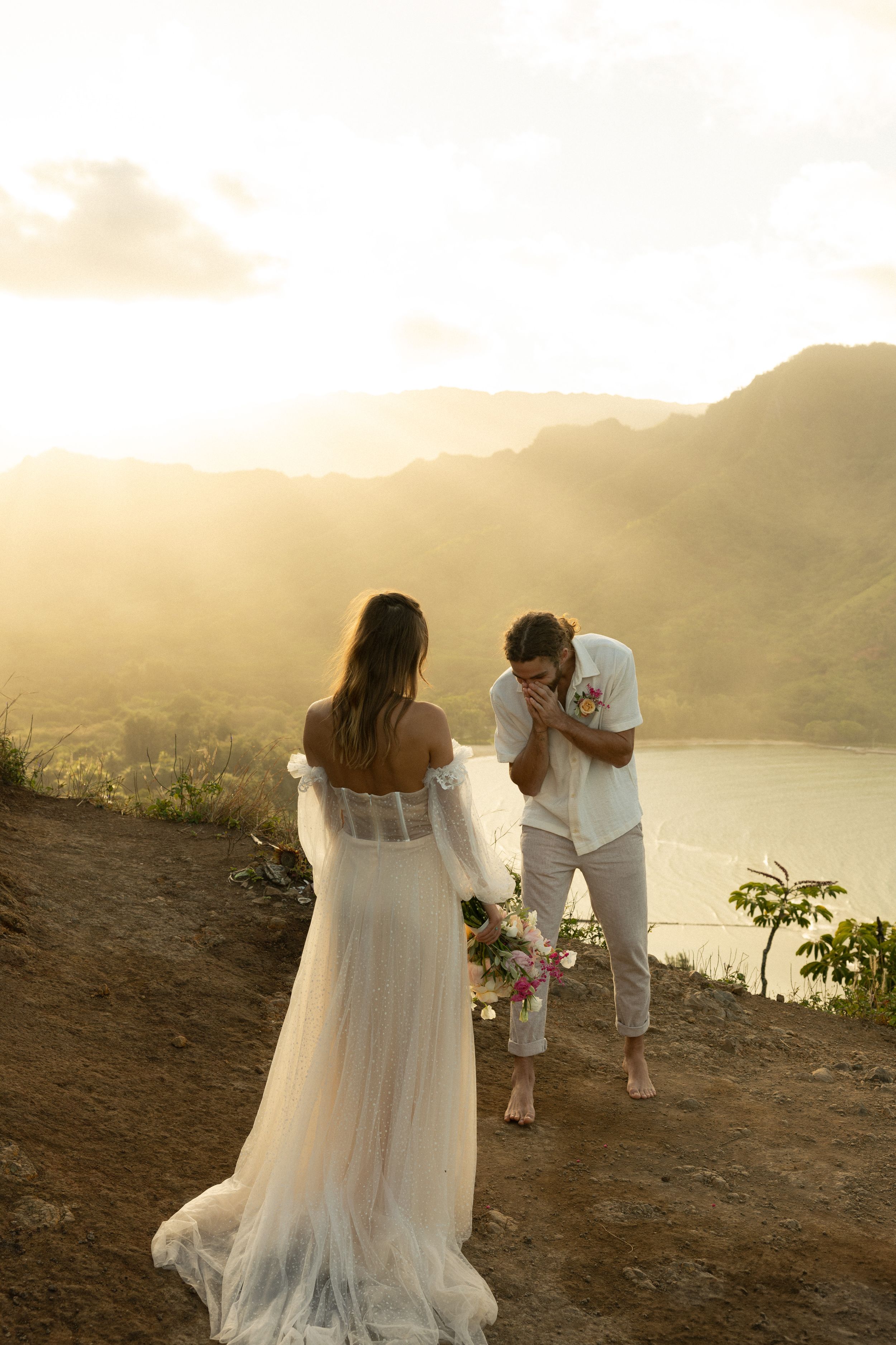 A couple shares a romantic moment at sunset overlooking mountains and water, with the bride in a flowing white dress.
