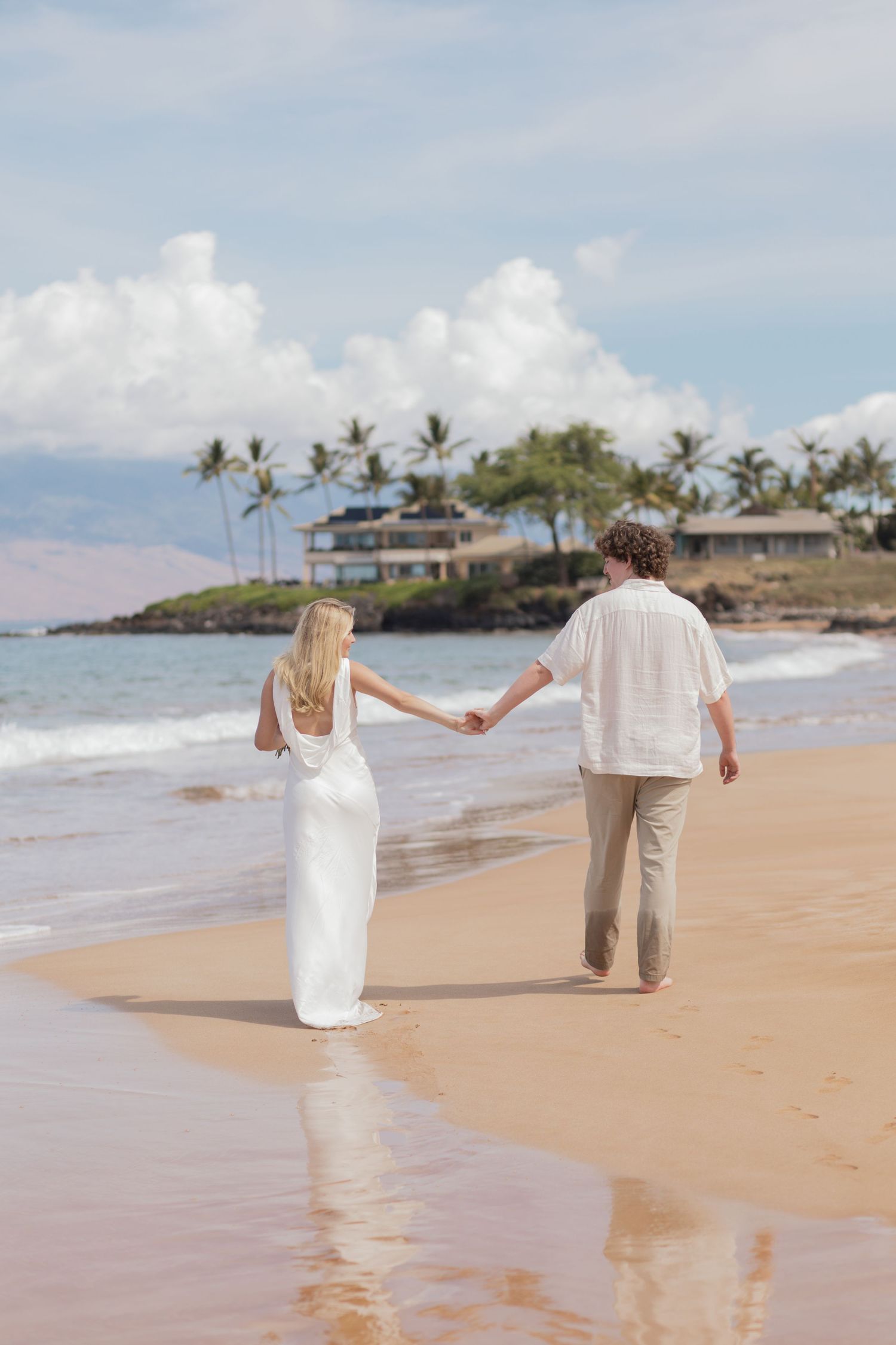 Two people holding hands walk along tropical shoreline in Hawaii with resort buildings visible in background.