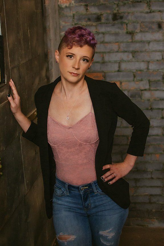 Professional photography of a figure in a black blazer, pink top and ripped jeans leaning against a brick wall.