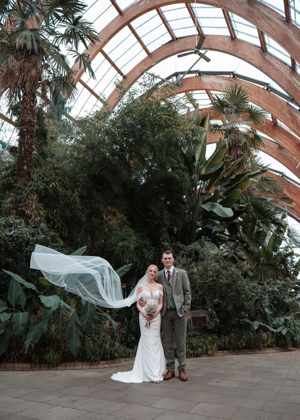 Peace Gardens Sheffield - Wedding couple posing in a beautiful greenhouse with arched glass ceiling and tropical plants.