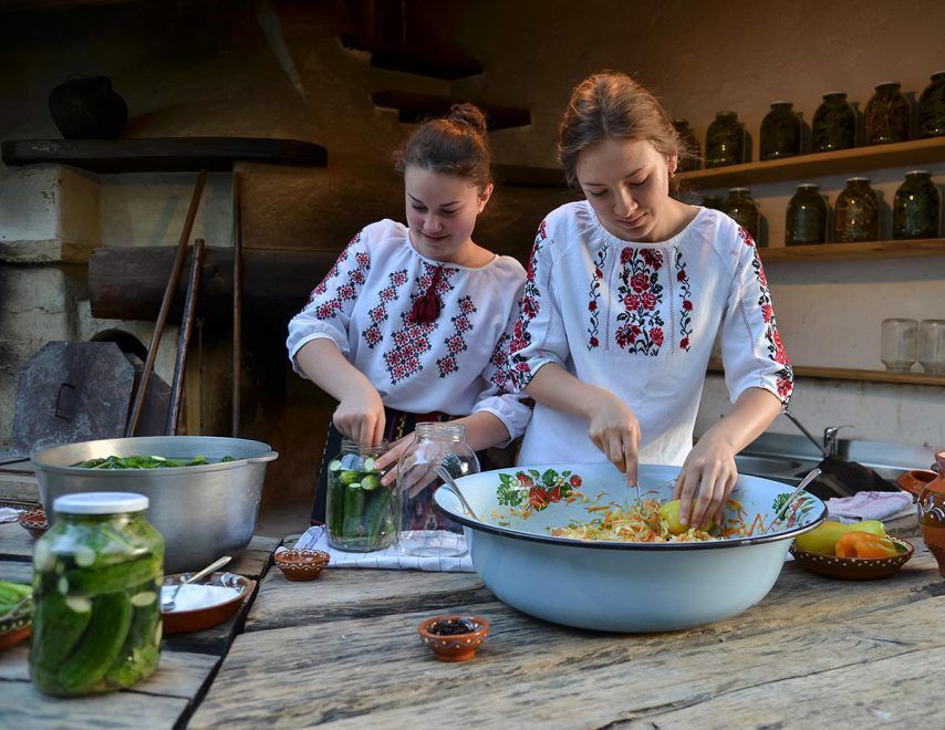 Two women in traditional embroidered blouses preparing food together at a rustic wooden table.