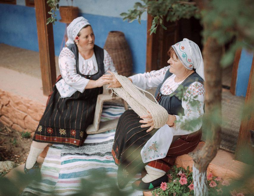 Two women in traditional folk costumes working with fabric outside a rustic blue building.