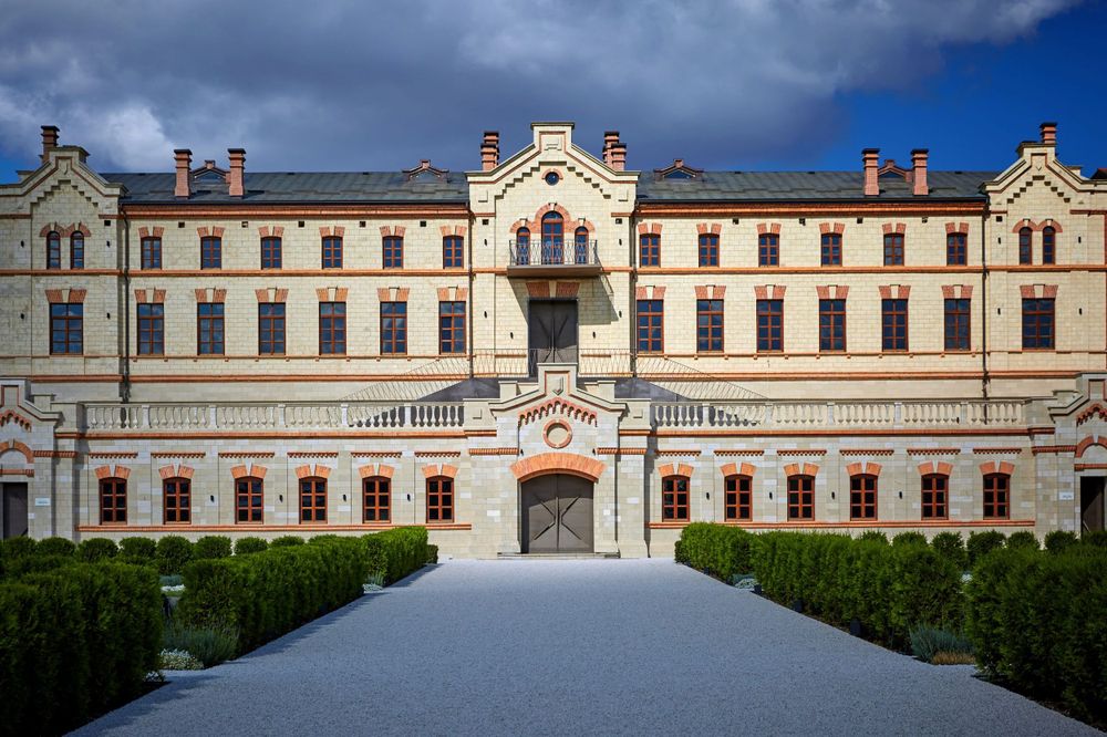 Grand historic palace facade with ornate architecture, manicured hedges, and cobblestone courtyard under dramatic sky.