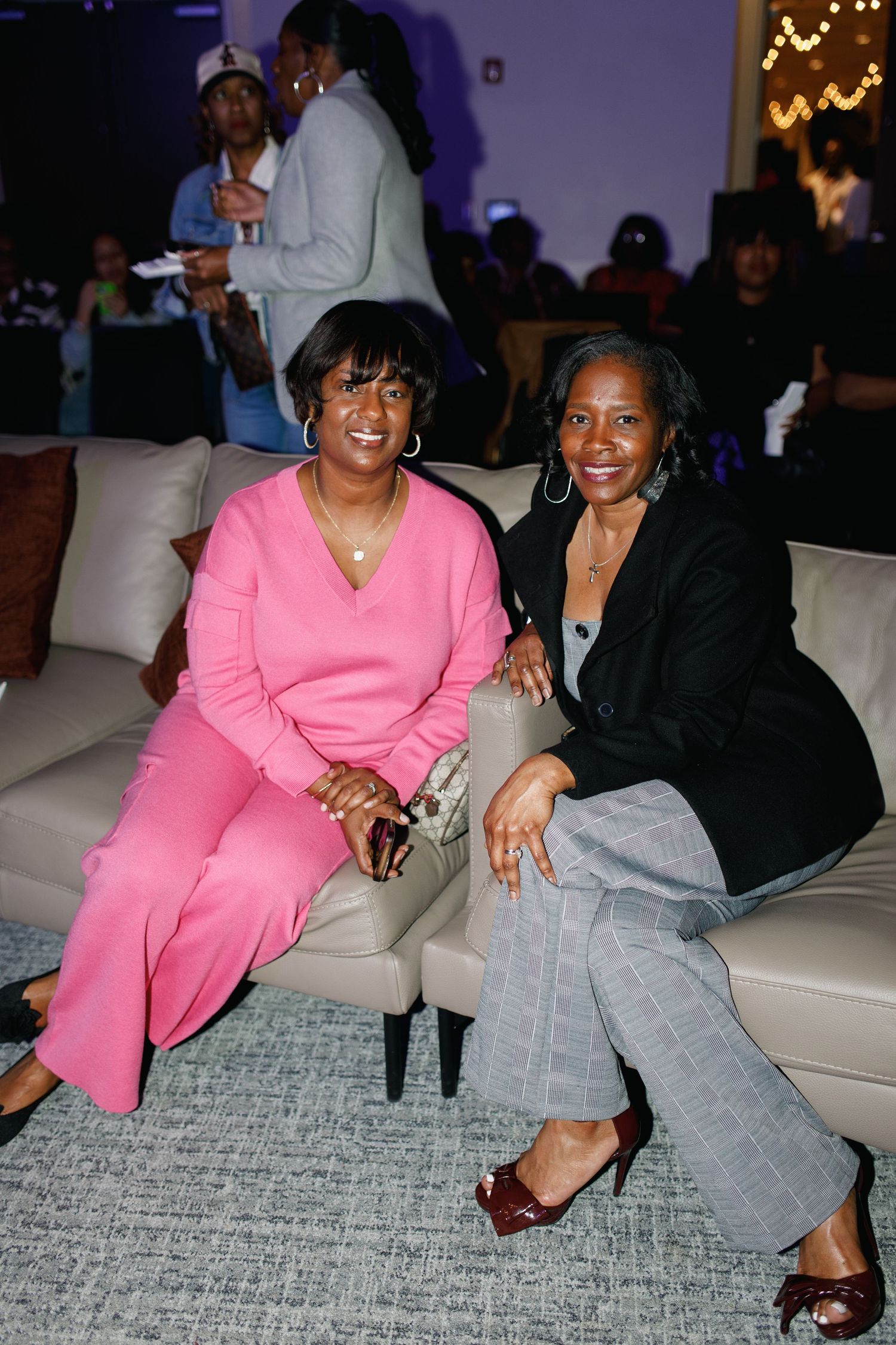 Two women smiling and posing together on a white sofa at an indoor event, one in pink and one in a black blazer.