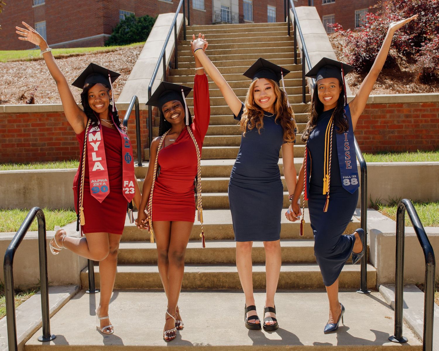 Four graduates in caps and gowns celebrating on campus steps with arms raised.