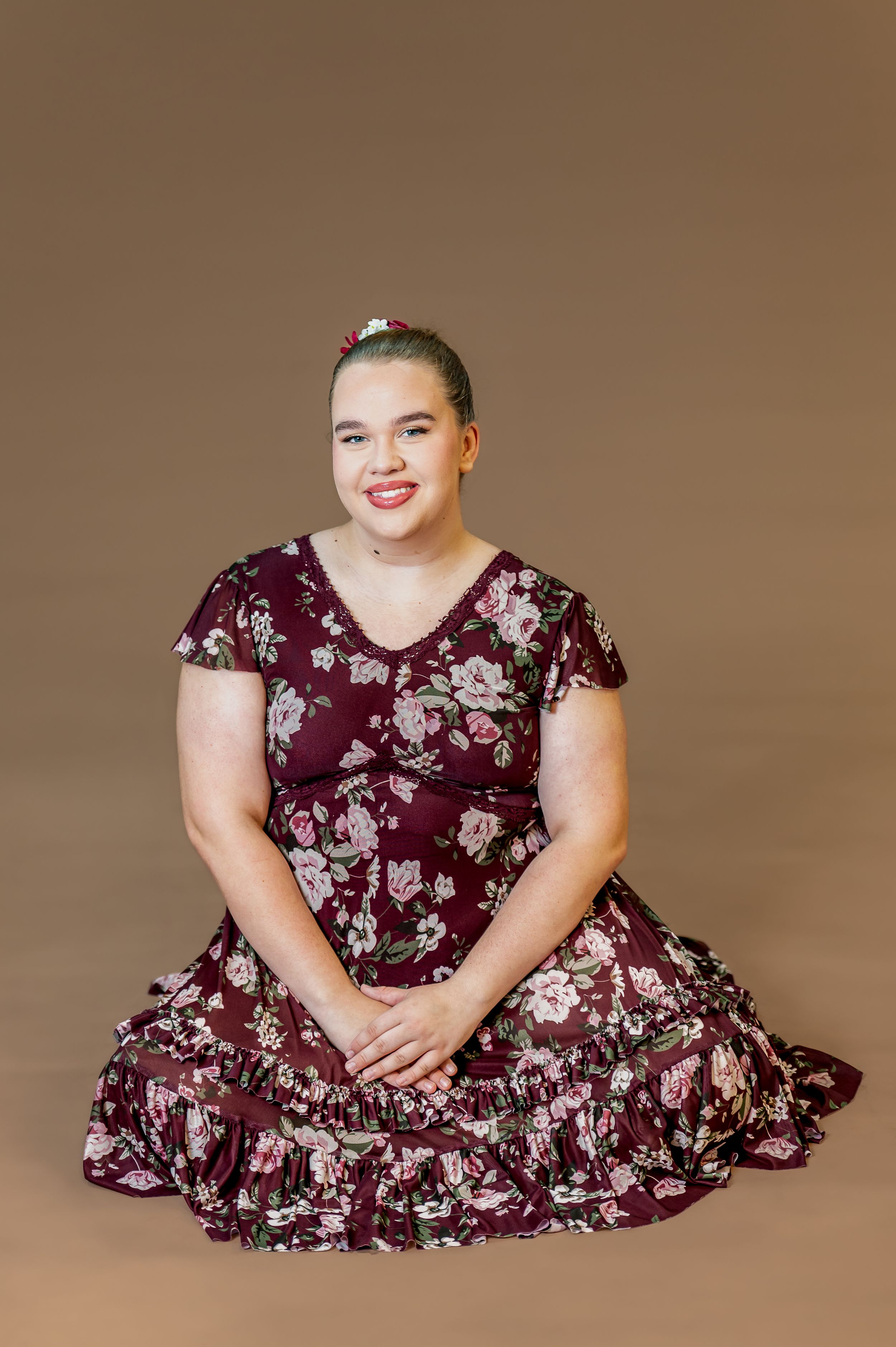Dancer poses seated in burgundy floral dress against brown background.