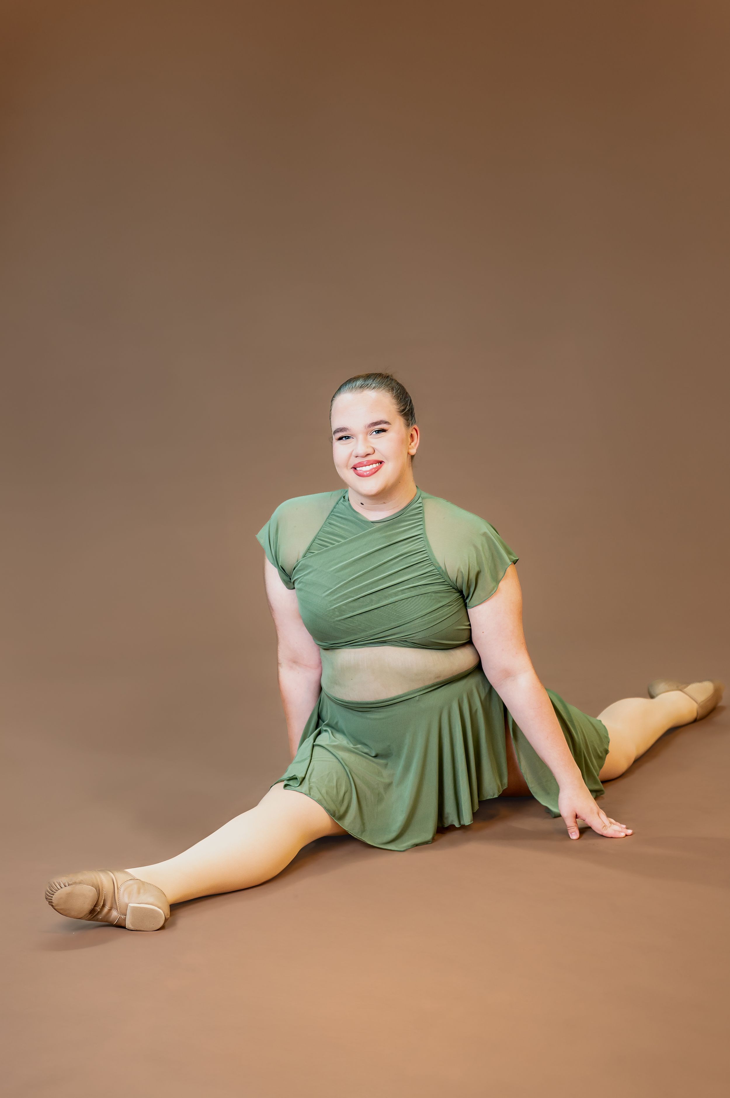 Dancer in green two-piece costume performs splits pose on brown studio backdrop.