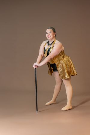Dancer in gold costume poses with dance cane against brown backdrop.