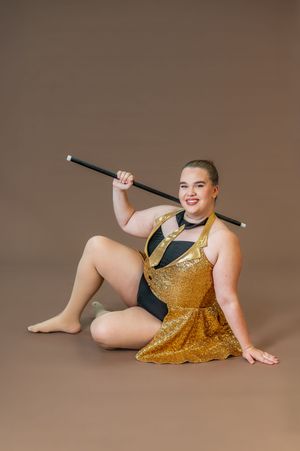 Dancer in sparkling gold and black costume poses with cane on brown backdrop.