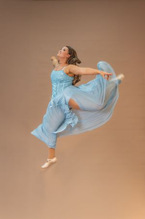 Dancer leaps gracefully in flowing light blue dress against brown background.