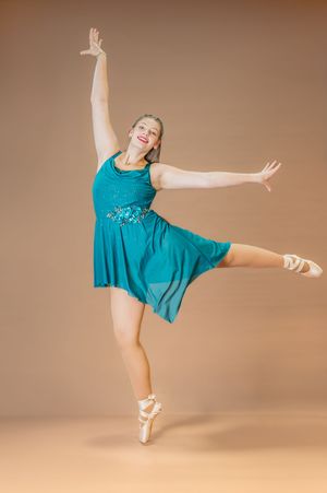 Dancer in flowing teal dress executes graceful arabesque on pointe shoes.