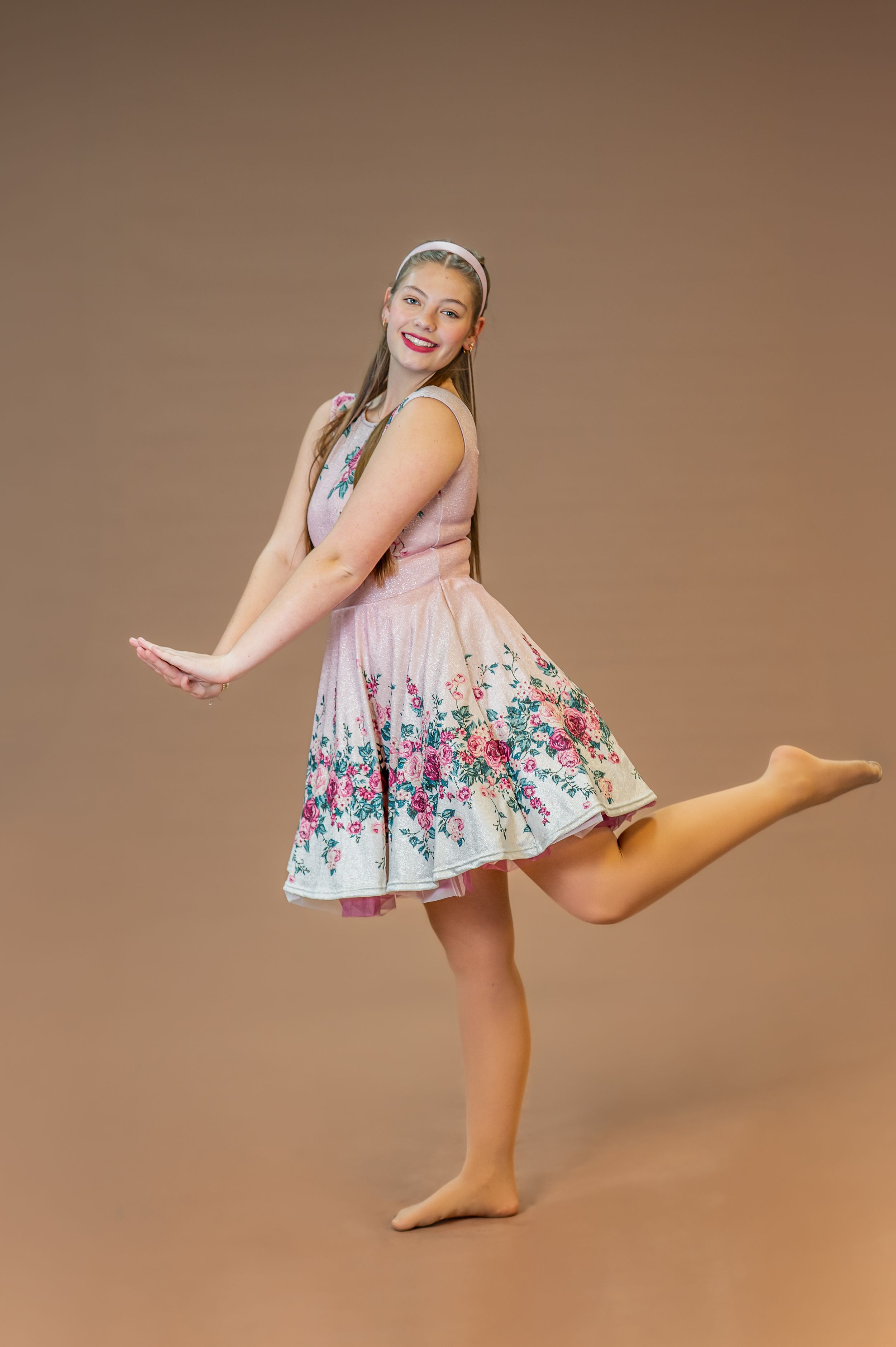 Dancer strikes playful pose in pink floral dress against beige backdrop.