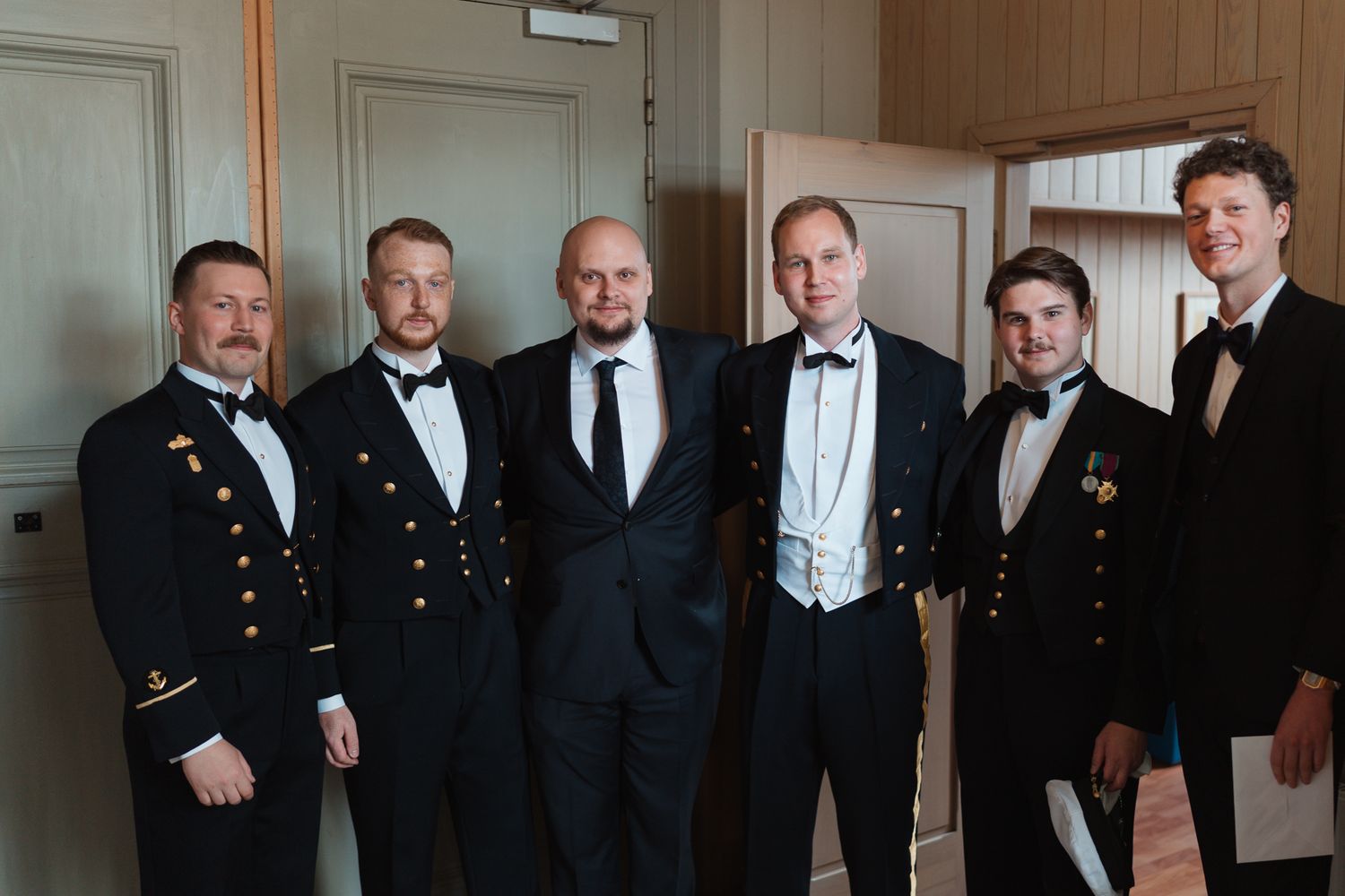 A group of six men in formal attire including tuxedos and military dress uniforms standing together indoors.
