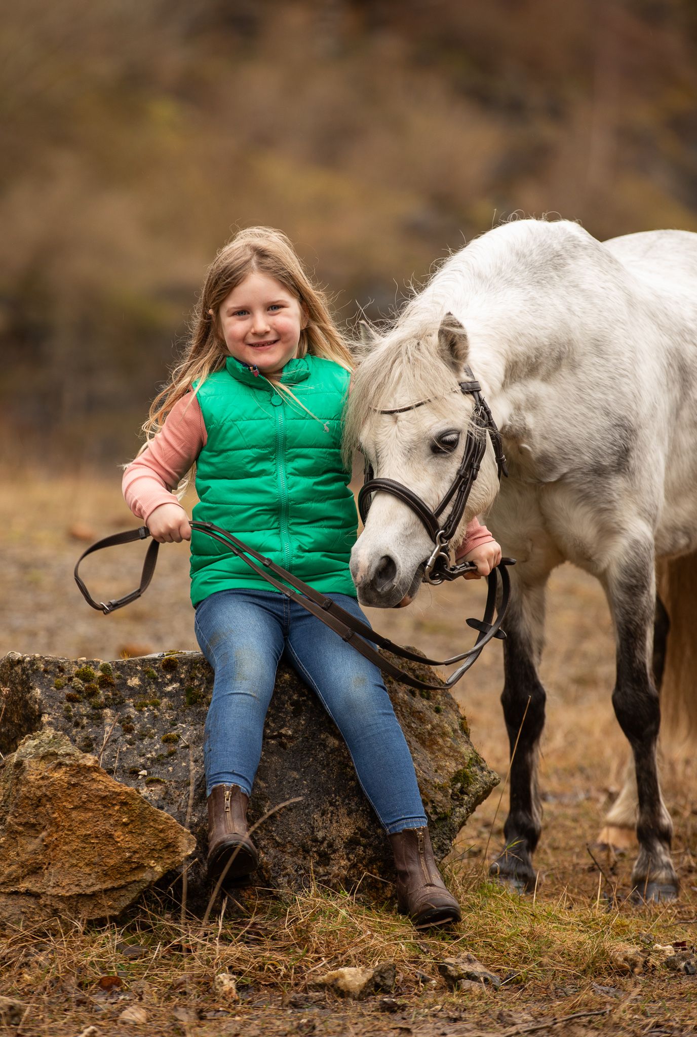 A white pony stands next to someone sitting on a rocky outcrop wearing a bright green vest and jeans in autumn scenery.