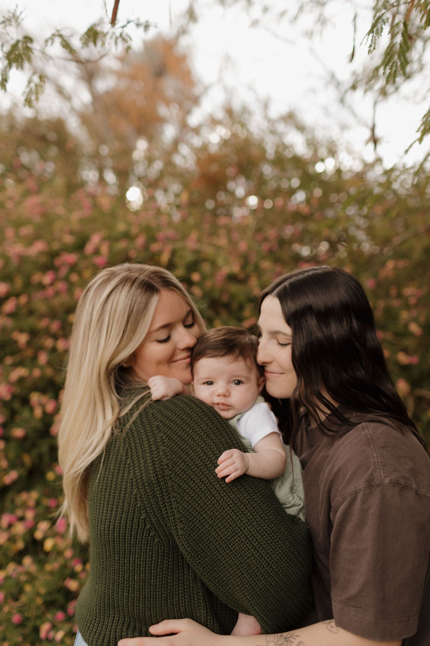 Two LGBT parents lovingly embrace their infant child during an outdoor autumn photoshoot.