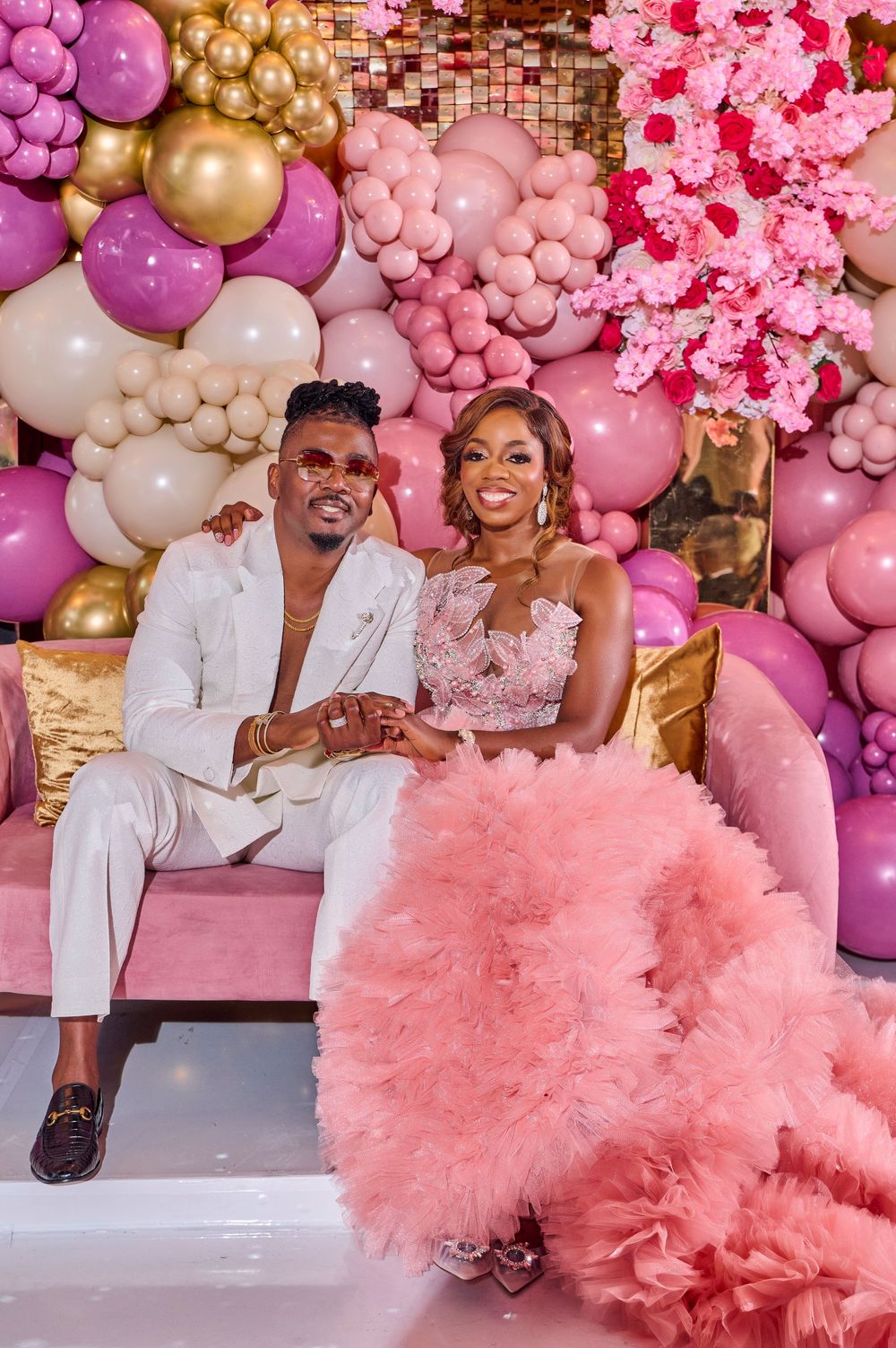 Romantic couple photo against pink and gold balloon backdrop with floral decorations.