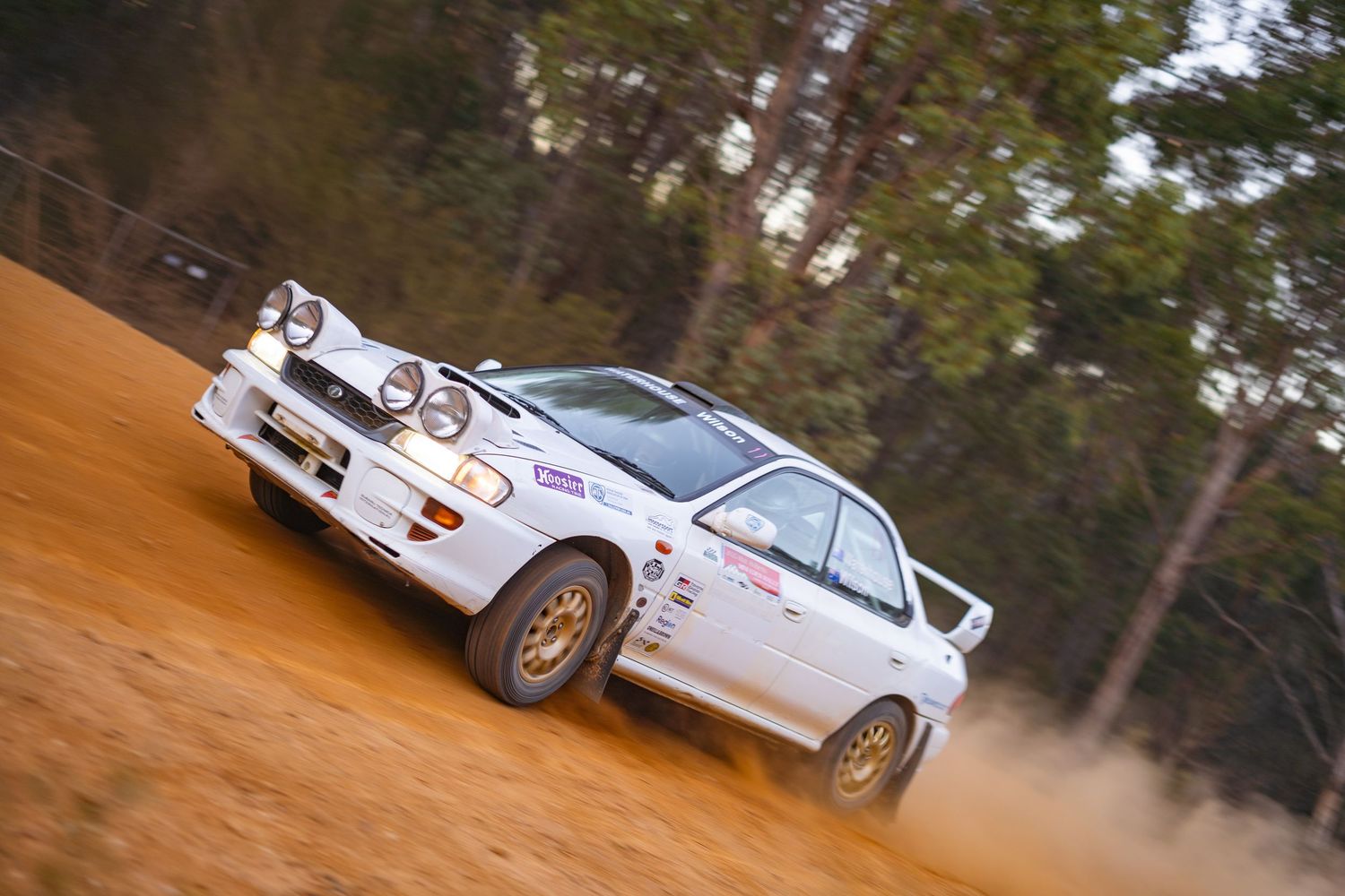 White Subaru WRX rally car kicks up dust while racing on dirt track through wooded area.