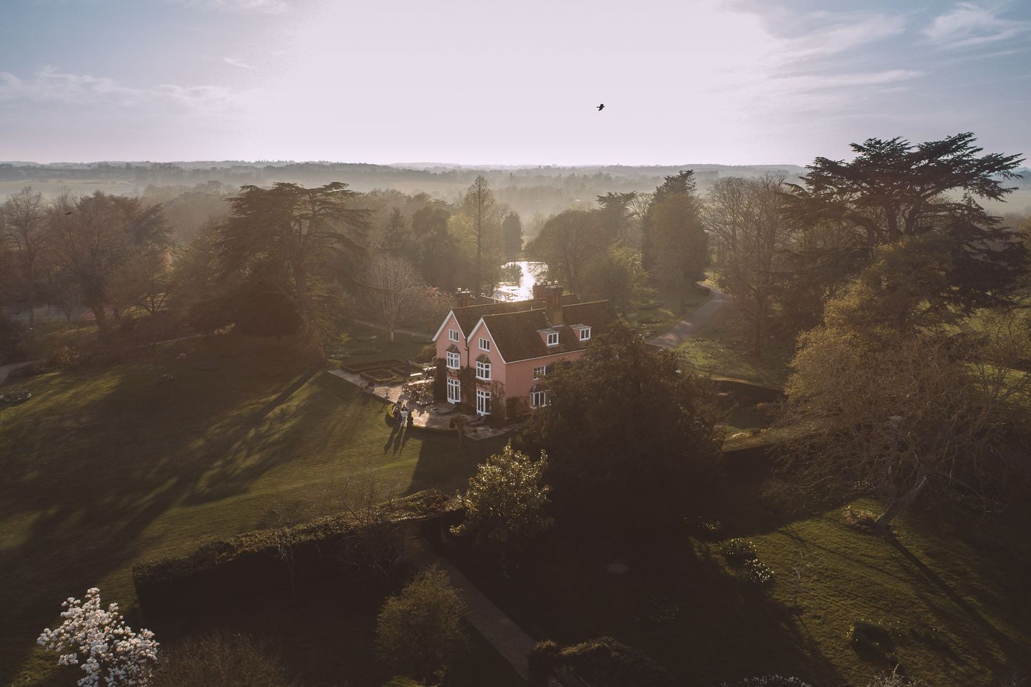 Aerial view of a charming Nedging Hall surrounded by lush green trees on a sunlit hilltop.