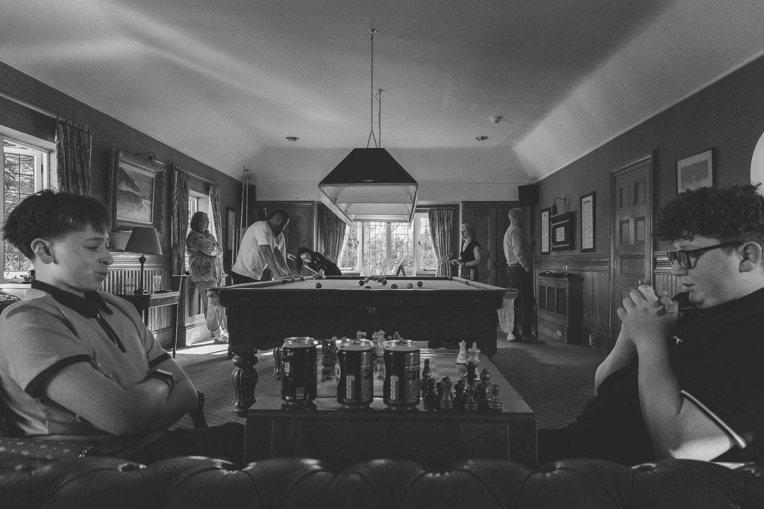Black and white image of people playing chess at a table in a dimly lit room with pendant lighting.