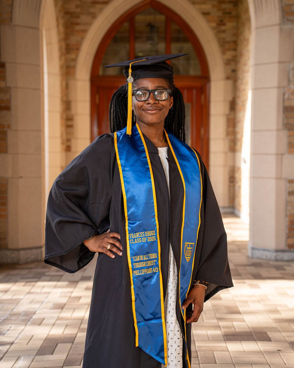 Graduate wearing a black cap and gown with blue and gold honors stoles stands in front of a stone archway.