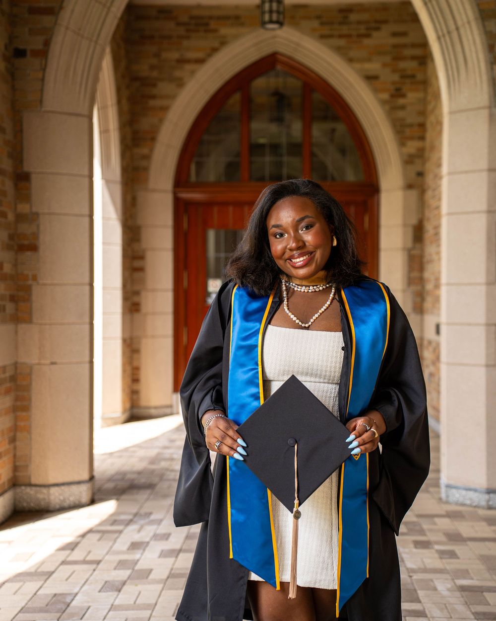 Graduate in traditional cap and gown with blue stole standing in arched stone corridor while holding graduation cap.