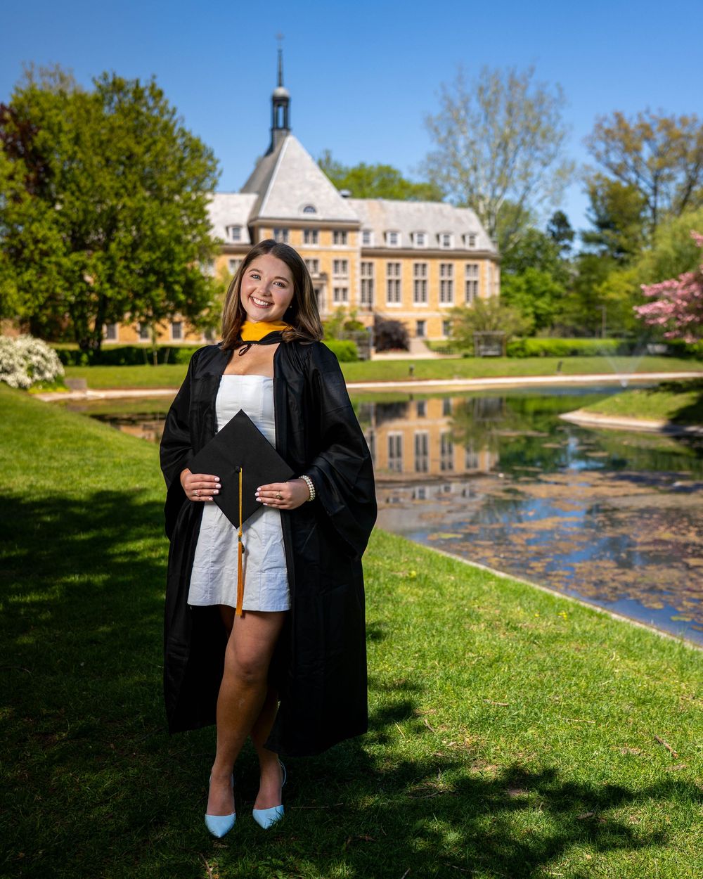 Graduate in academic regalia poses on lawn with historic campus building and reflecting pond in background.