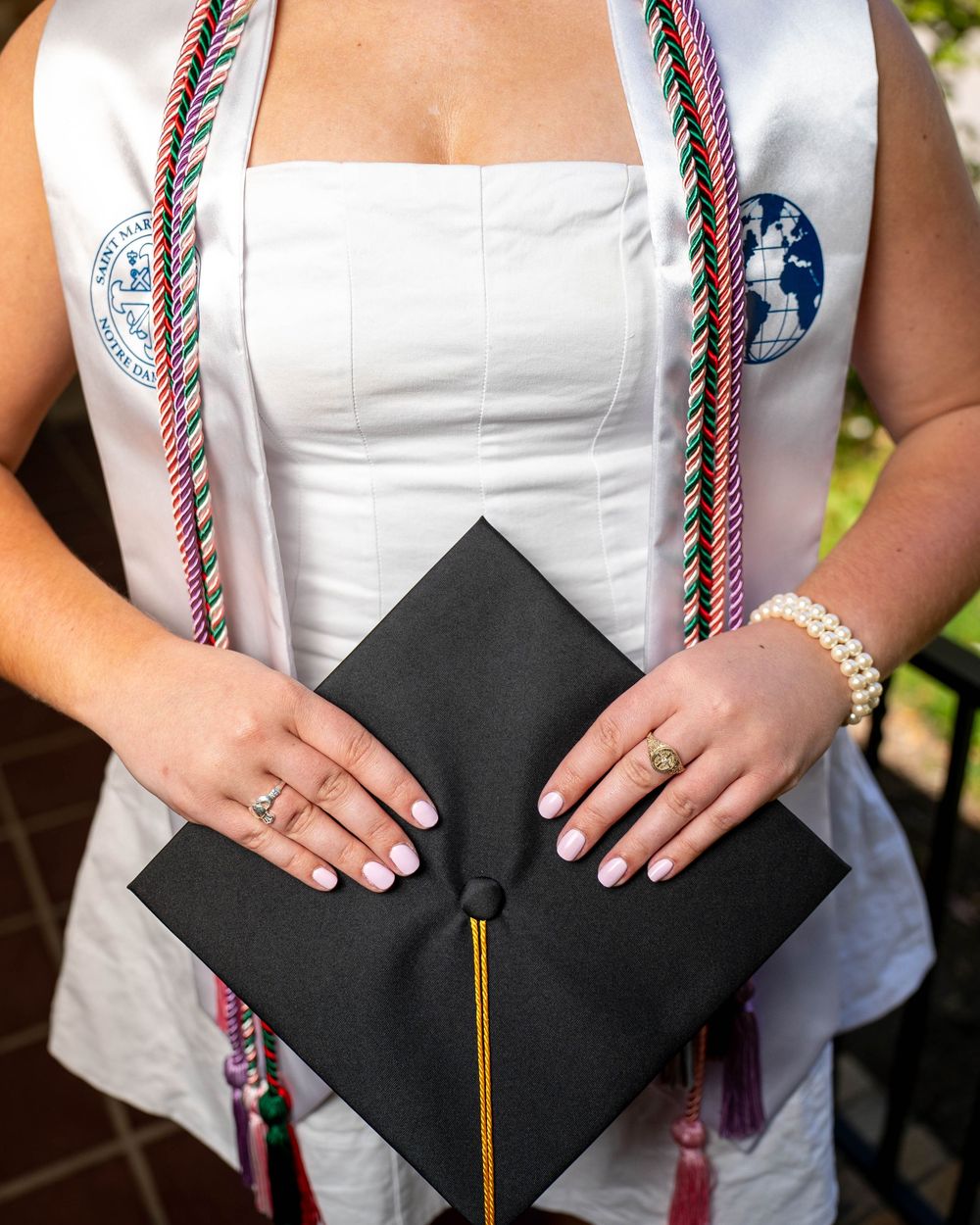Graduate holding a black mortarboard graduation cap while wearing pearls and traditional graduation attire.