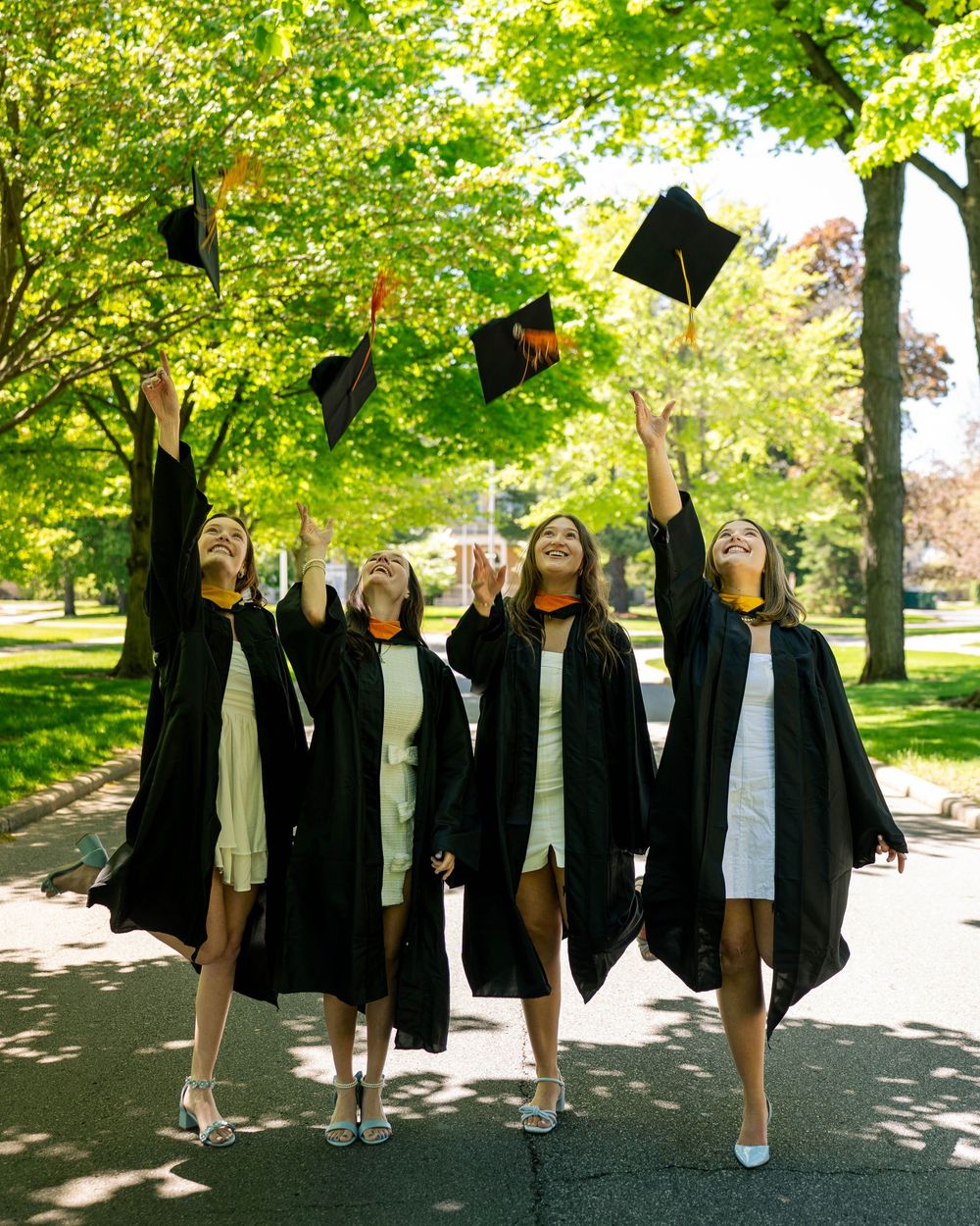 Graduates in black caps and gowns toss their mortarboards into the air while celebrating on a tree-lined path.