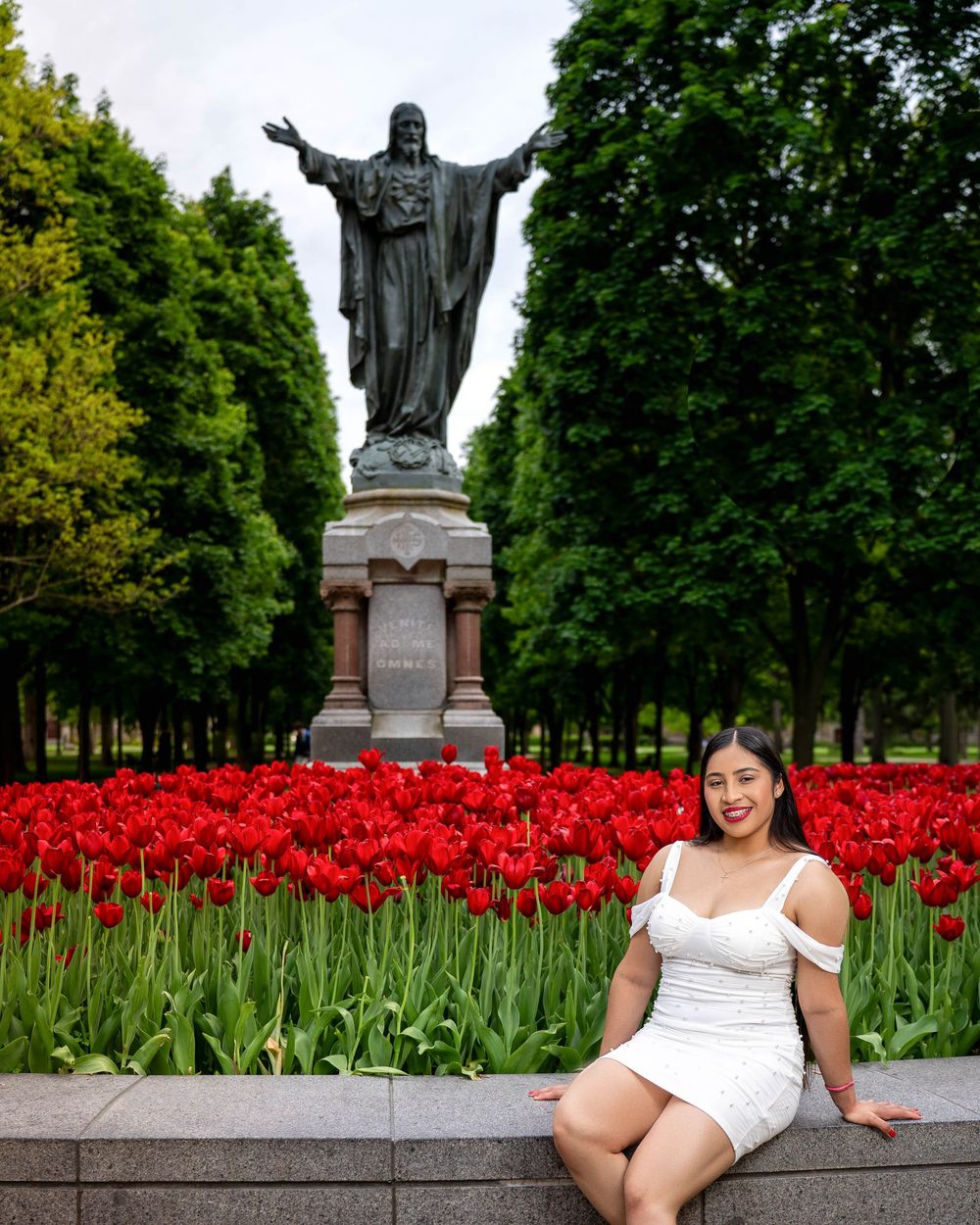 A large religious statue stands among vibrant red tulips in a serene garden setting.
