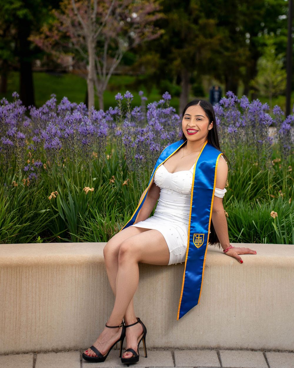 Graduate in white dress and blue sash sits on stone ledge with purple wildflowers blooming in garden background.