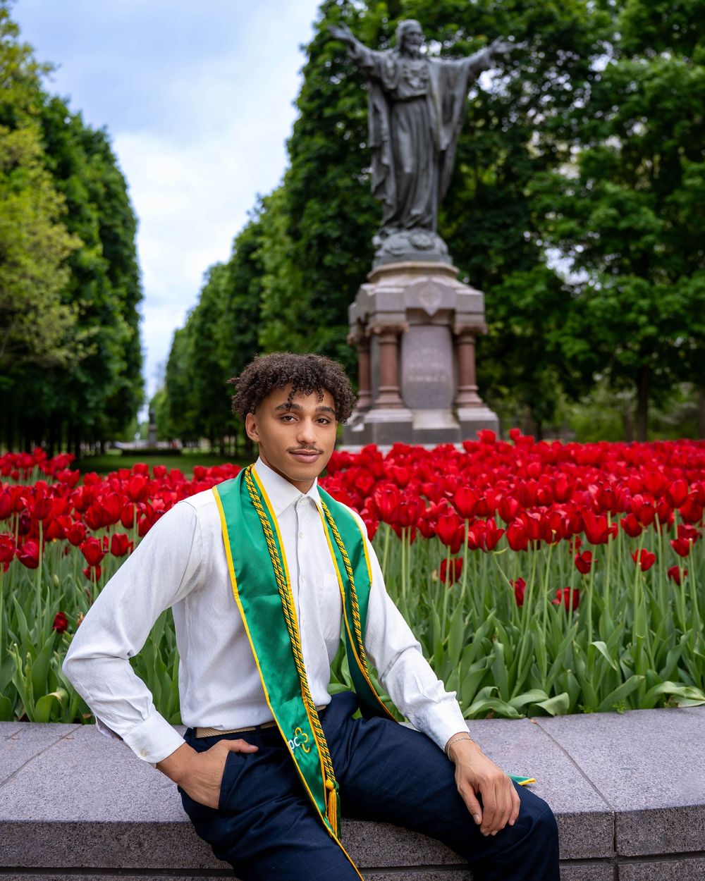 Graduate wearing green sash poses for photo in front of red tulip garden and statue on sunny spring day.