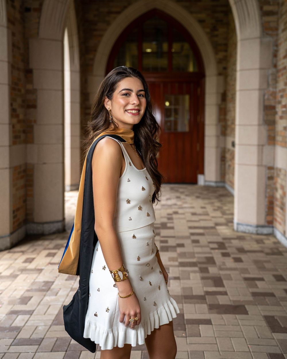 Student wearing a white summer dress smiles in a historic archway corridor with tiled flooring.