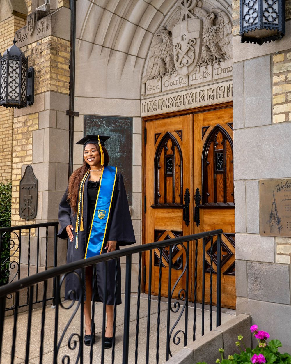 Graduate poses in cap and gown in front of ornate wooden church doors with a blue graduation sash.