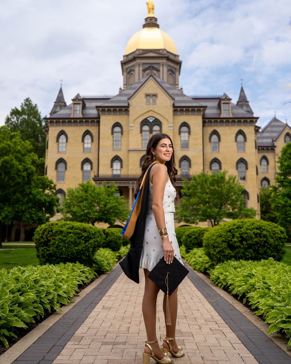 A graduate in cap and gown stands in front of Notre Dame's iconic Golden Dome building on a sunny spring day.