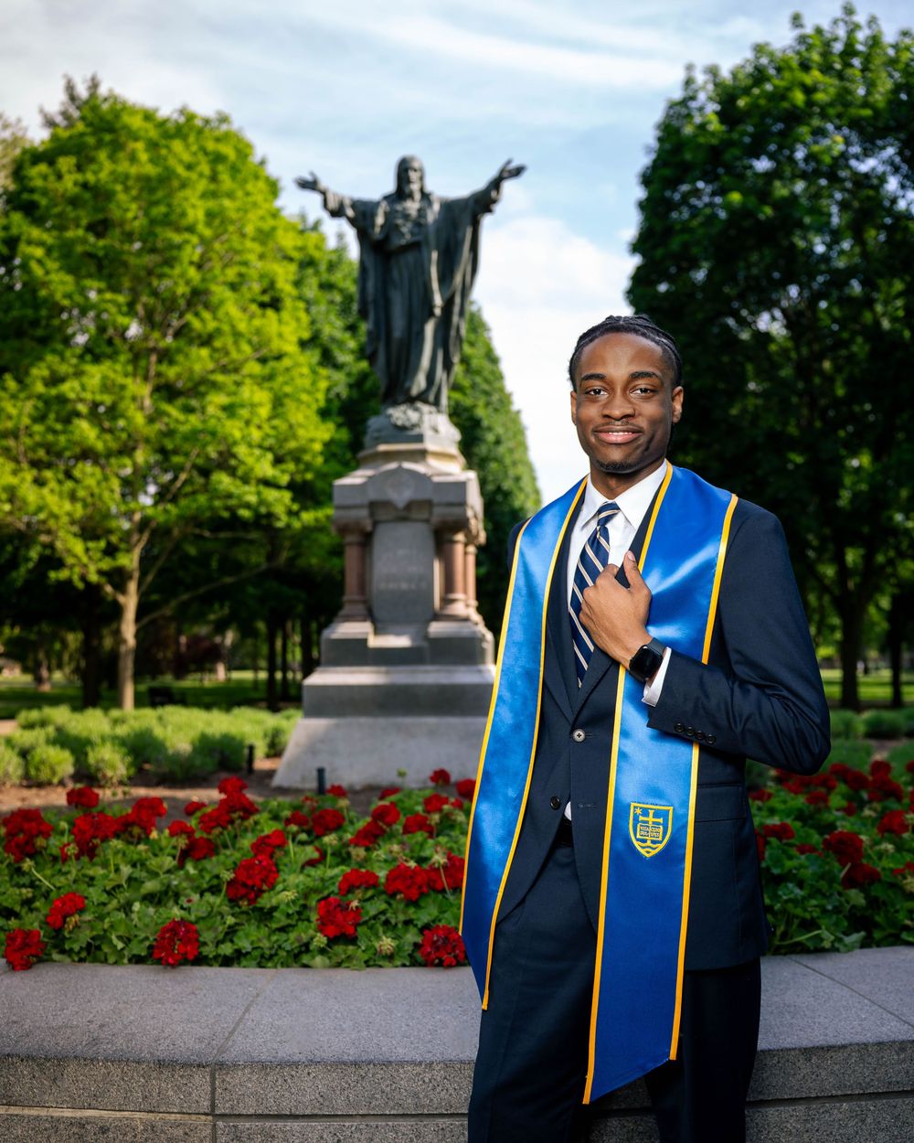 Graduate in academic regalia poses near statue and red flower garden on college campus.
