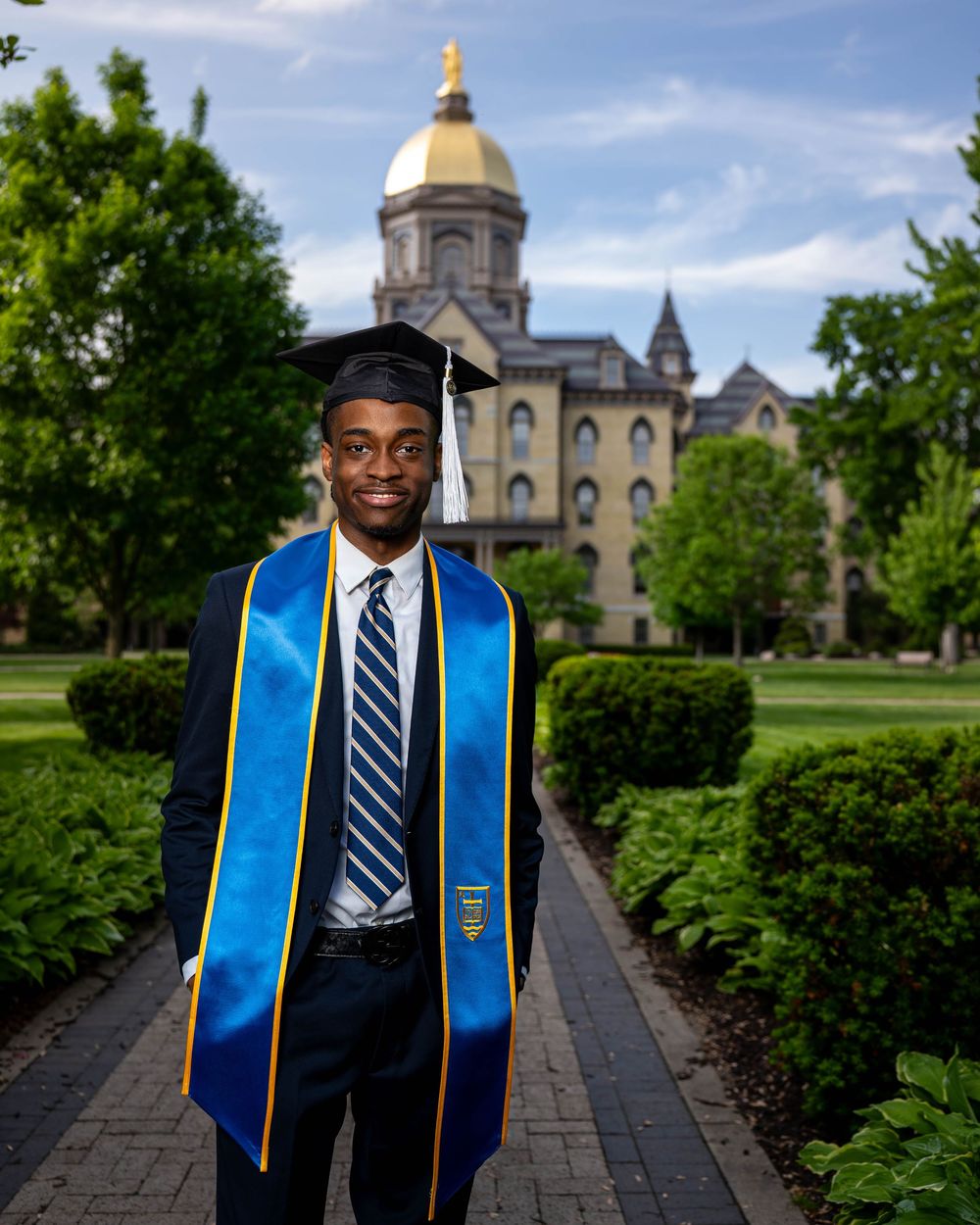A graduate poses in academic regalia on a pathway with the iconic Notre Dame Golden Dome building visible in the background.